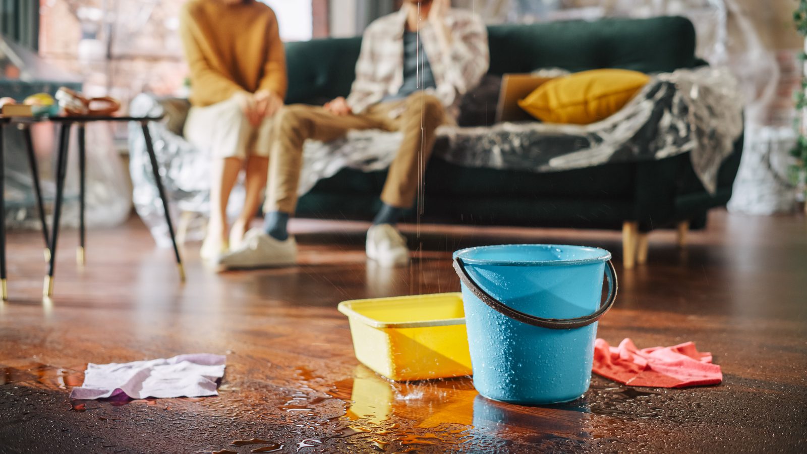Couple sitting on a plastic-covered sofa with buckets catching a water leak in their rental
